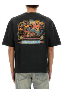 Amiri men’s black t-shirt featuring a vibrant vintage-style tropical hideaway graphic with palm trees and architecture
