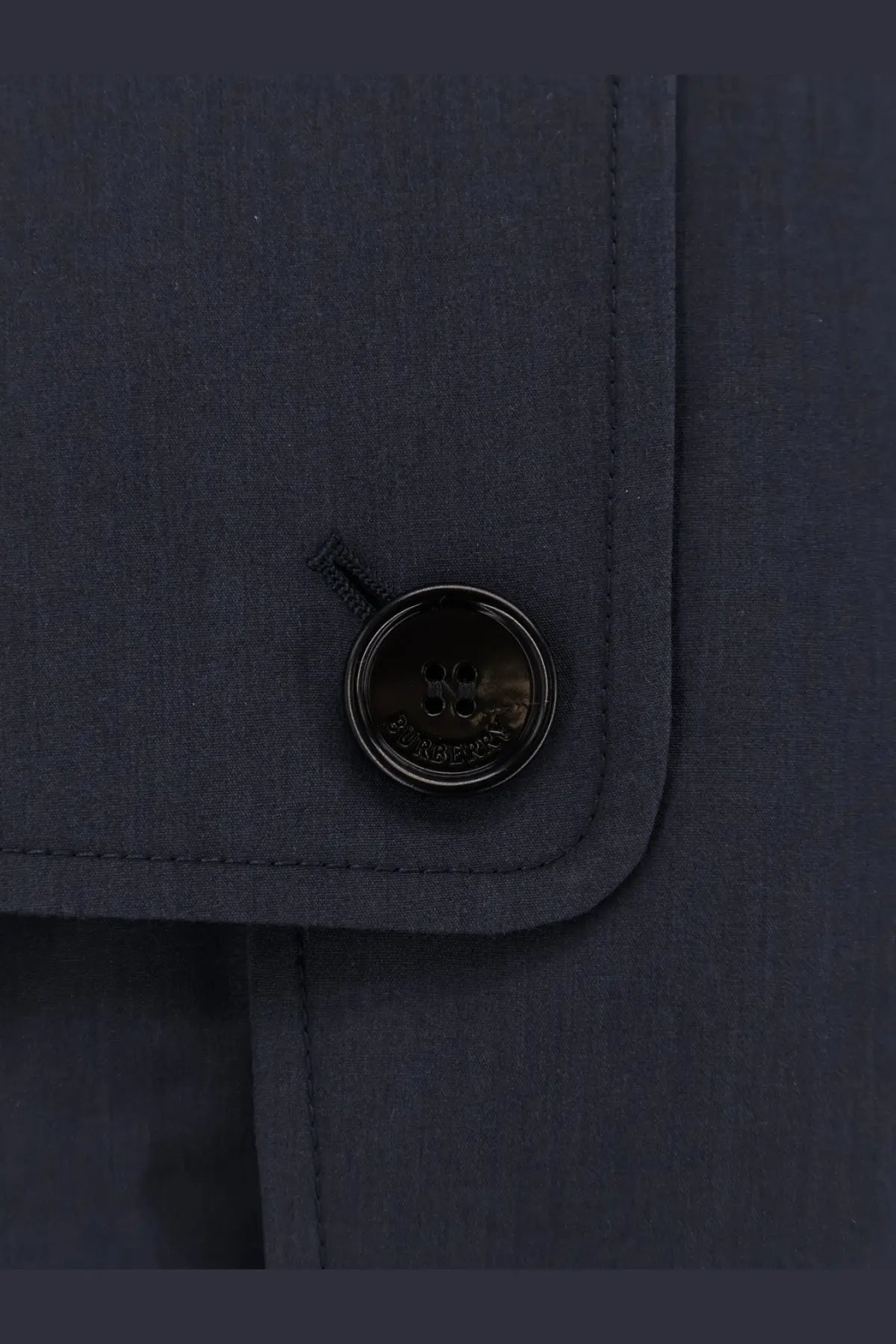 Black circular button with embossed Burberry logo on dark navy cotton blend fabric of Burberry men’s Highbury trench