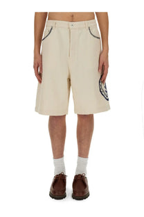 Beige Drôle de Monsieur men’s cotton bermuda shorts with black embroidered pockets and circular logo patch on leg