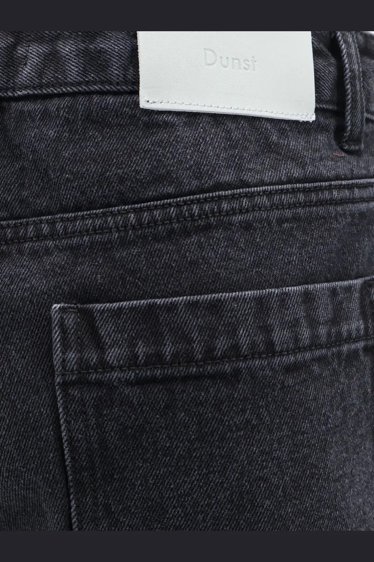 Back view of dunst men’s black denim bermuda shorts with dark-wash fabric and white leather brand label on waistband