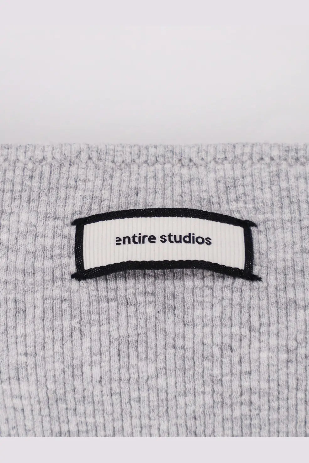 A white ribbed label with entire studios text sewn onto a heather gray textured knit fabric of a women’s ribbed cotton tank top pebble