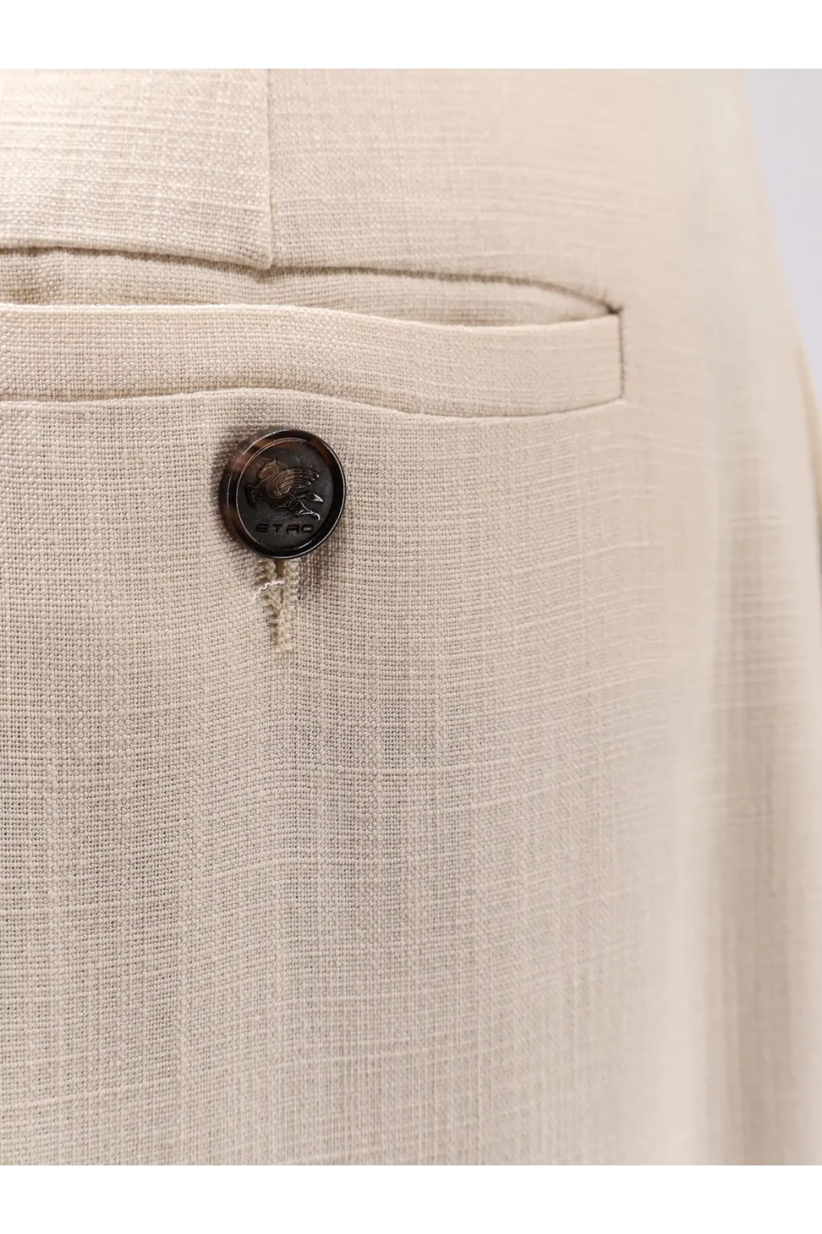 A dark circular button with embossed design on textured beige linen fabric of etro women’s white viscose trousers