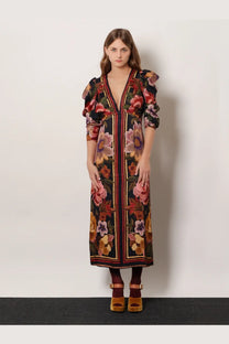A woman wears a farm rio women’s viscose long dress black with vibrant floral print and gold trim along the front opening