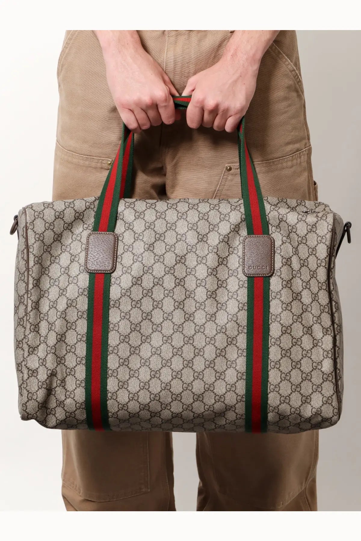 Beige Gucci men’s GG supreme canvas duffel bag with red and green striped handles for travel use