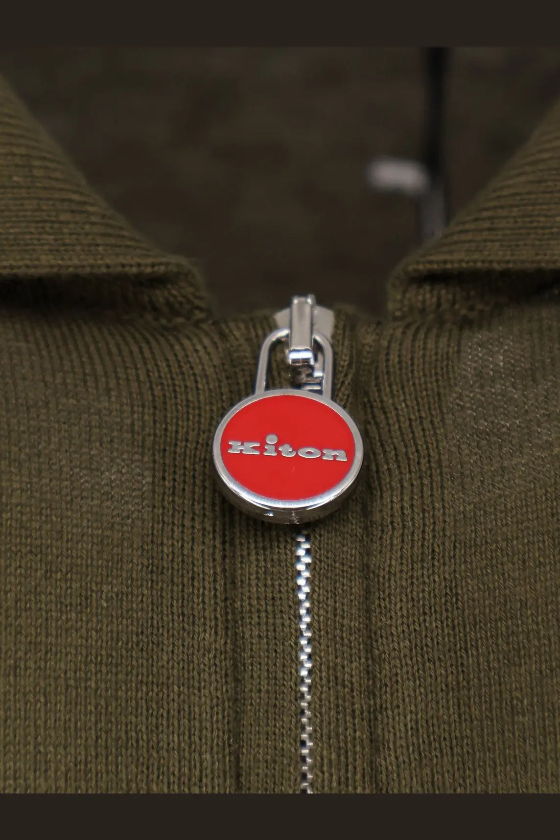 Red circular kiton zipper pull with silver trim and white brand lettering on kiton men’s green cotton polo shirt