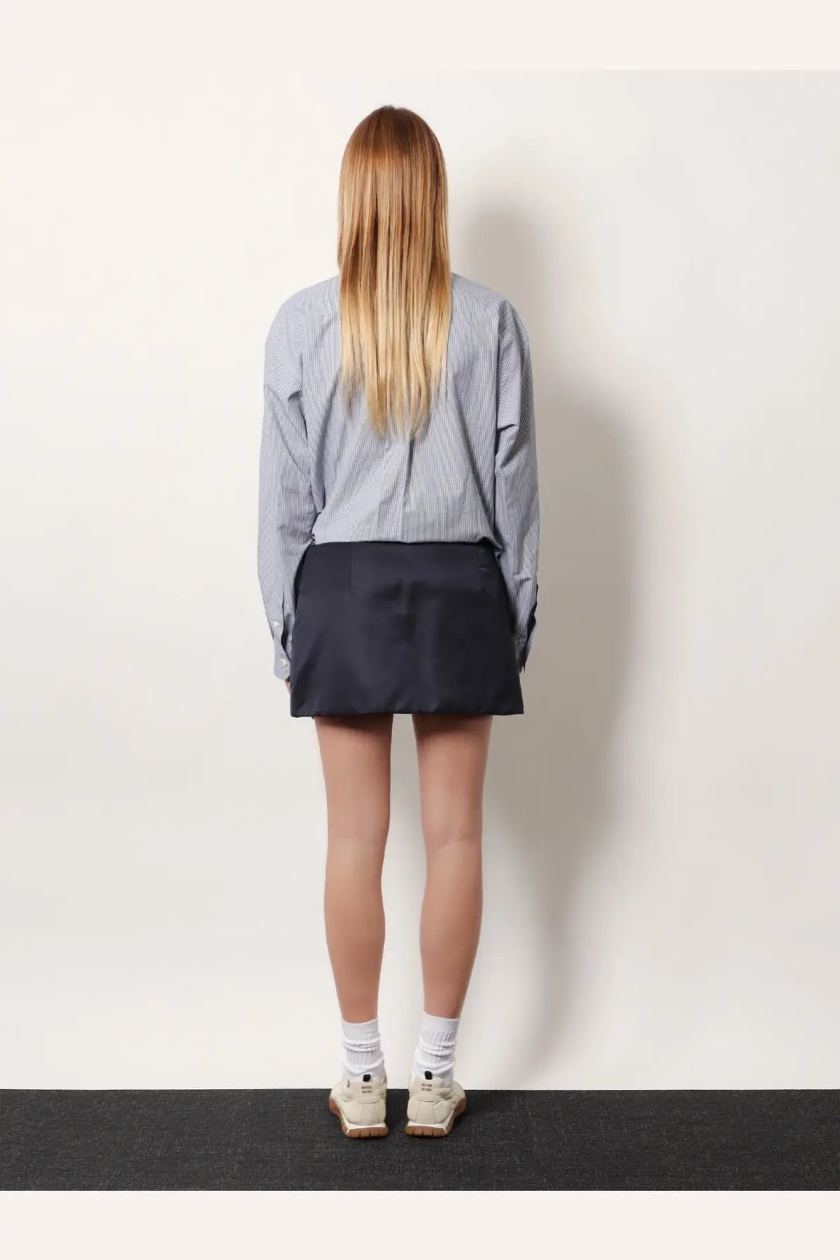 A woman wears a dark navy miu miu mini skirt made from recycled material paired with a blue and white striped shirt