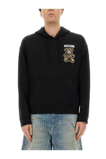 Black Moschino men’s hoodie featuring a brown embroidered teddy bear patch with the brand name above it