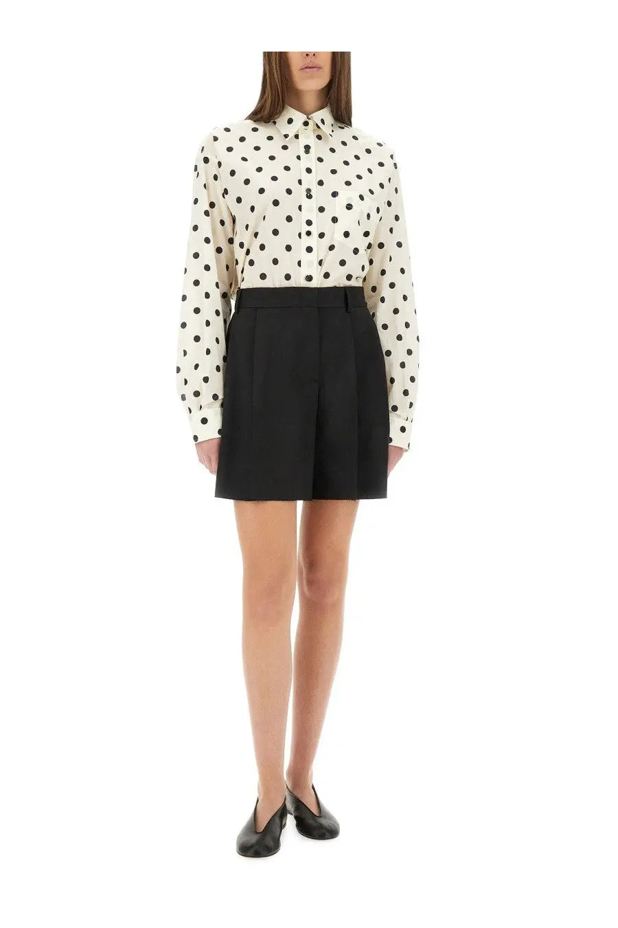 A woman wearing moschino women’s asymmetrical black wool high-waisted pleated shorts with a cream polka dot blouse