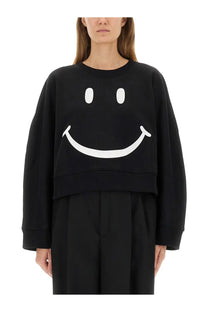 Moschino women’s black cropped sweatshirt made of soft fabric featuring a large white smiley face graphic on front