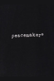 Black fabric tote bag with white peacemaker logo embroidered in sans serif font for oamc peacemaker men’s cotton shirt black