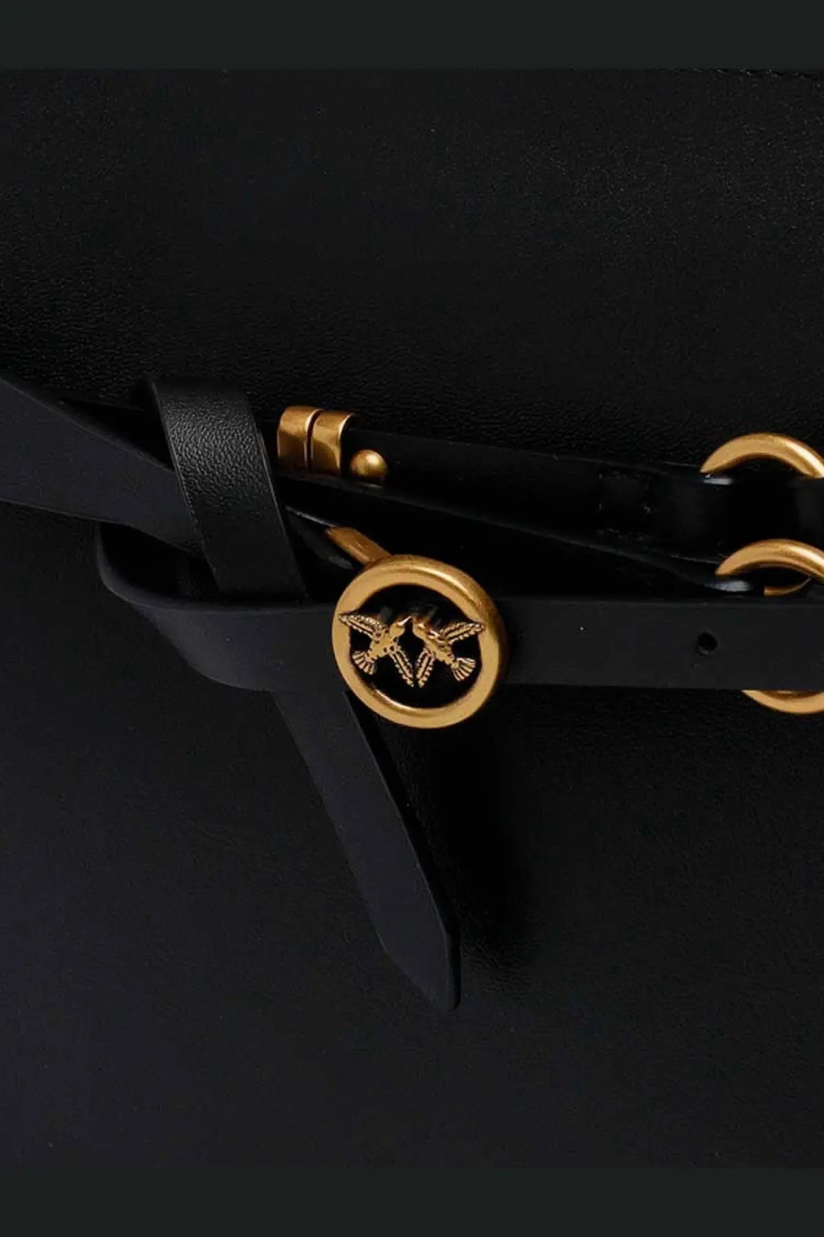 Black leather strap with gold-tone circular buckle featuring embossed bird design on pinko women’s leather belt bag crossbody bag