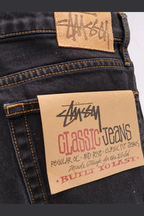 Dark-wash denim jeans with tan leather brand patch and paper label featuring stussy logo in black script back view