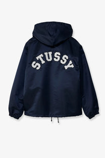 Back view of stussy men’s navy blue nylon coach jacket with white arched lettering across the back