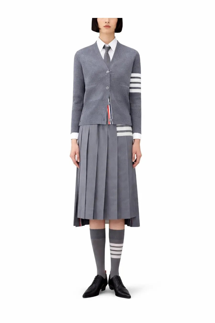 Thom Browne women’s gray tweed neck cardigan with three white sleeve stripes worn over white shirt and pleated skirt