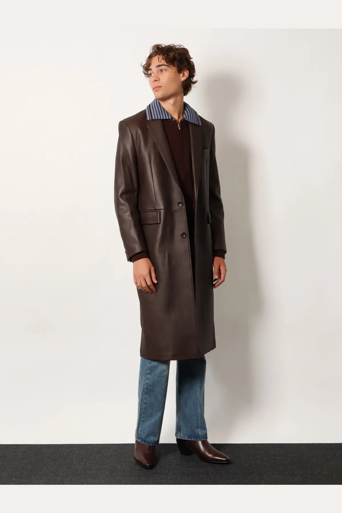 Tonywack unisex flared leg jeans in blue paired with a sleek dark brown leather overcoat with a blue and white striped collar