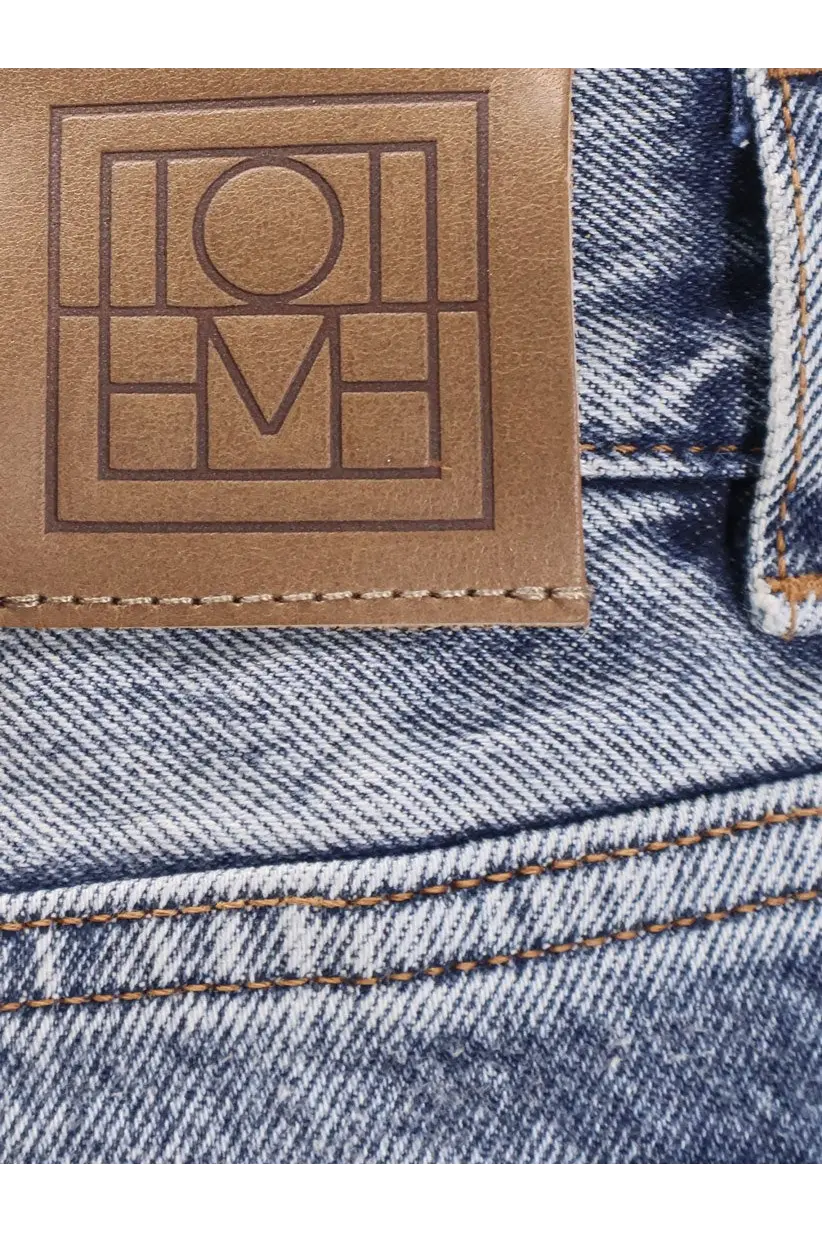 Brown leather patch with embossed loewe logo on the back of grey Toteme women’s cotton jeans on blue denim fabric