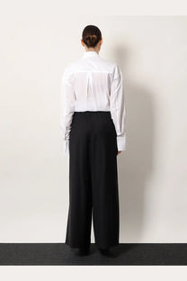 Back view of a white button-down shirt with subtle vertical pleats and long sleeves paired with toteme women’s black wool trousers