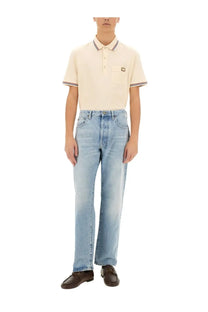 Cream-colored short-sleeved polo shirt with navy white striped collar and cuffs and black logo patch on chest pocket