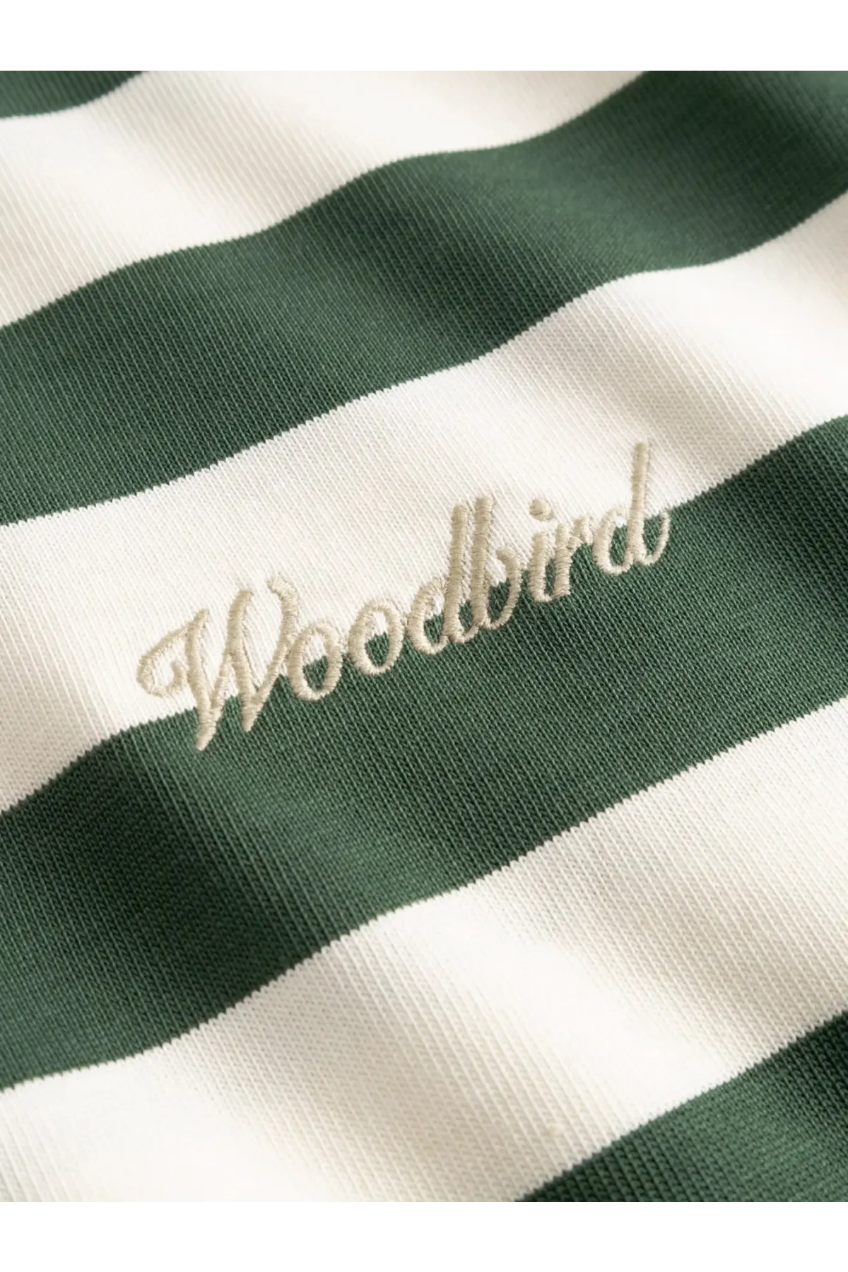A cream-colored embroidered woodbird logo on a green and white striped organic cotton woodbird men’s tee